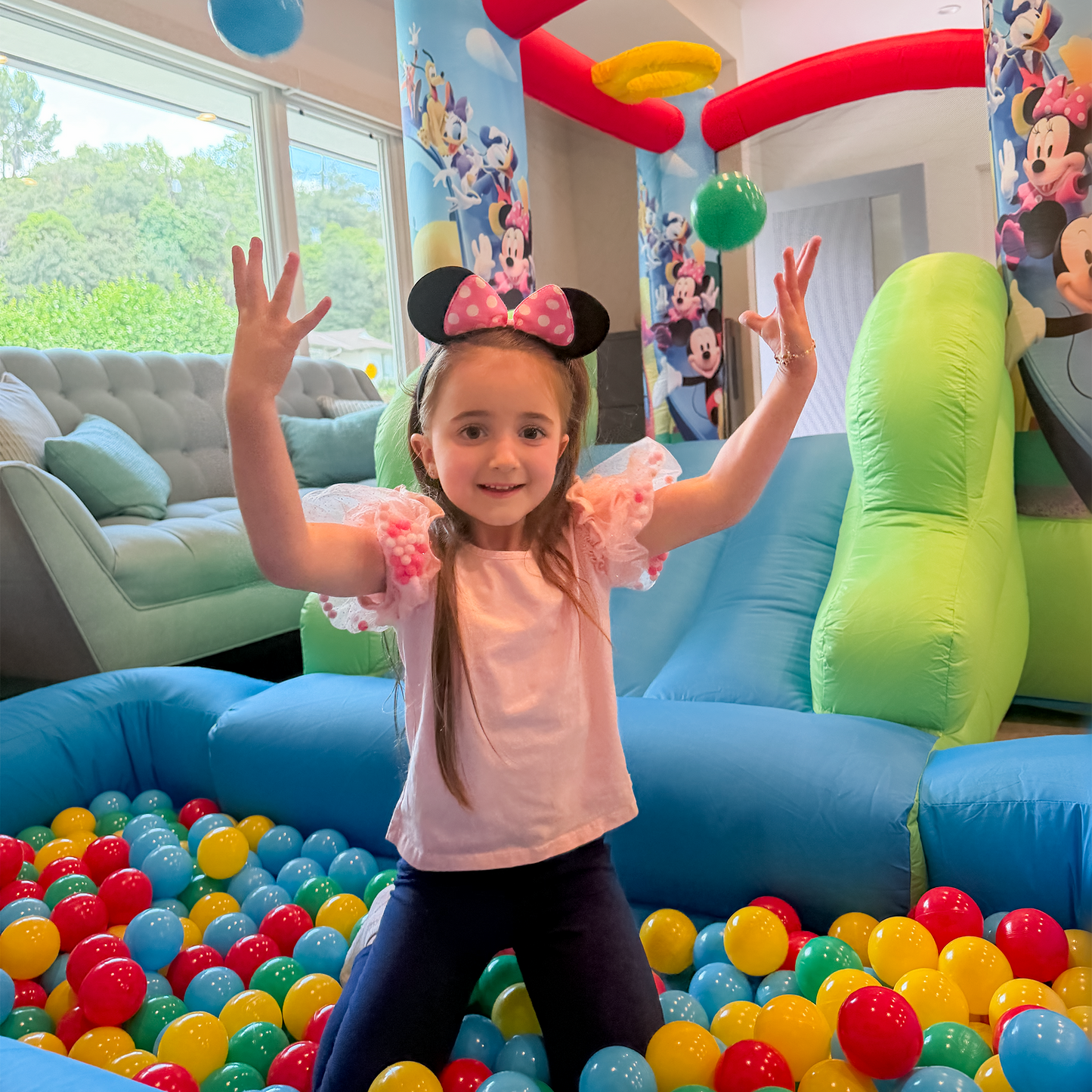 Child in a ball pit wearing Minnie Mouse ears in a playroom with colorful balloons and decorations.